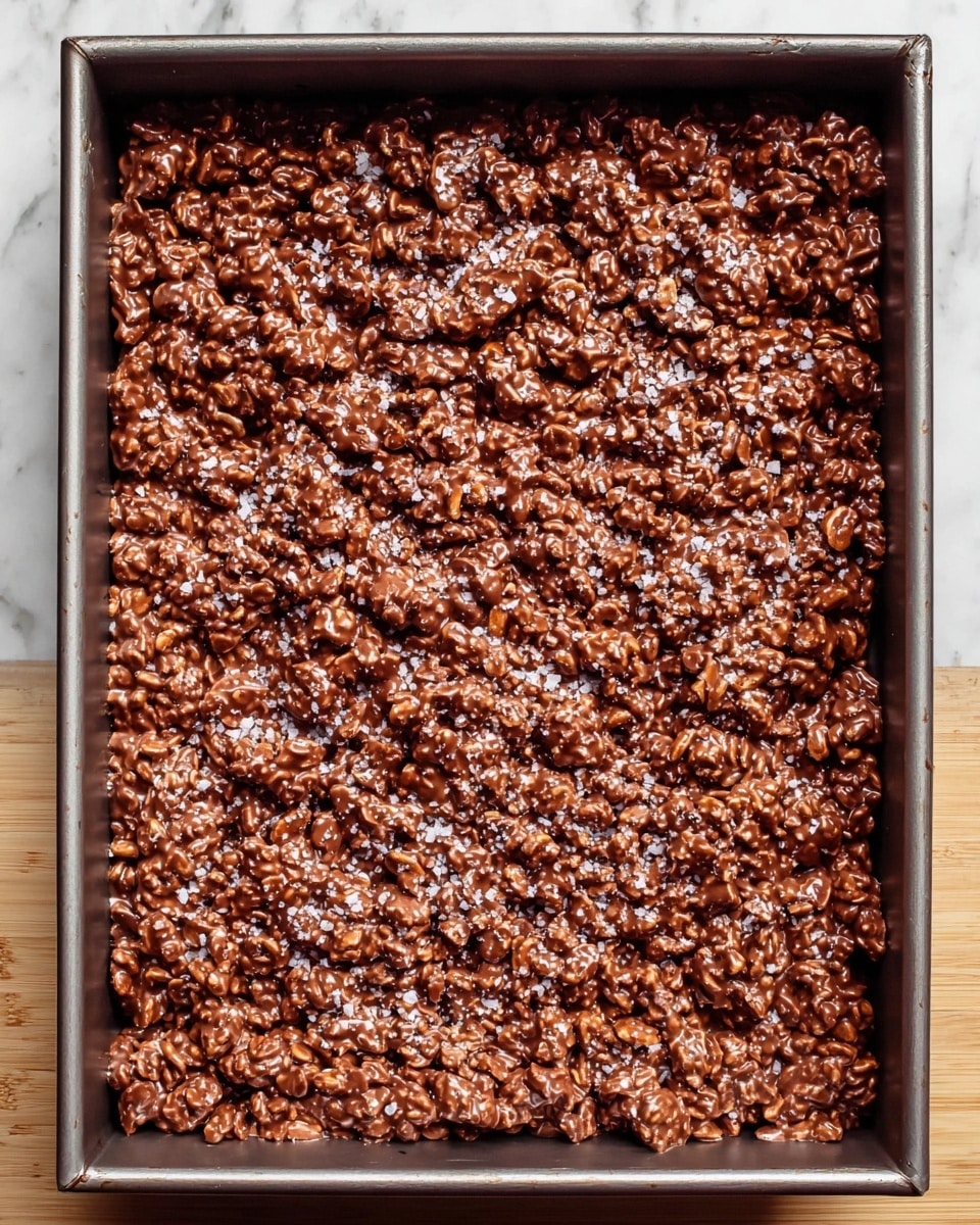 The image shows a single thick layer of uneven, chunky chocolate clusters spread tightly in a rectangular metal baking pan. The chocolate coating has a glossy, shiny surface with visible textures of nuts or cereal bits mixed inside. Some small sprinklings of white salt flakes are scattered on top, adding a slight contrast to the deep brown color. The pan is set on a white marbled surface. photo taken with an iphone --ar 4:5 --v 7