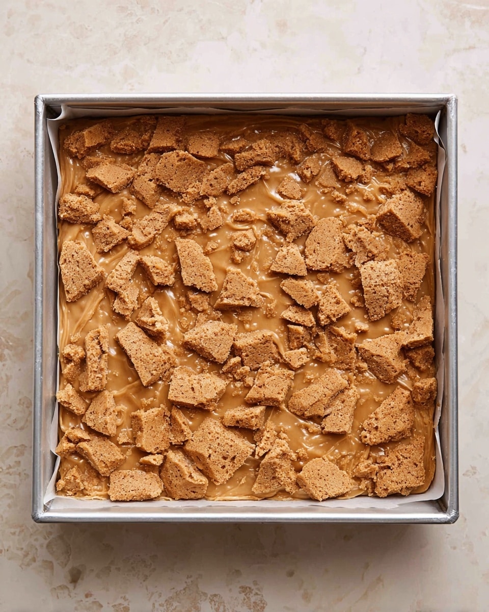 The image shows a square metal baking pan lined with parchment paper, filled with a thick, light brown cookie dough base. On top, there are scattered broken pieces of golden brown cookies, some large and some small, spread evenly across the surface. The cookie pieces have a rough texture and are embedded slightly into the dough, which has some shiny melted spots around them. The baking pan rests on a white marbled surface. photo taken with an iphone --ar 4:5 --v 7