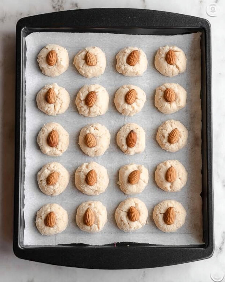 A black baking tray lined with white parchment paper holds 20 small, round dollops of light beige dough evenly spaced in a 4x5 grid. Each dough mound has a soft, slightly rough texture and is topped with a single whole almond placed in the center. The background is a white marbled surface. photo taken with an iphone --ar 4:5 --v 7