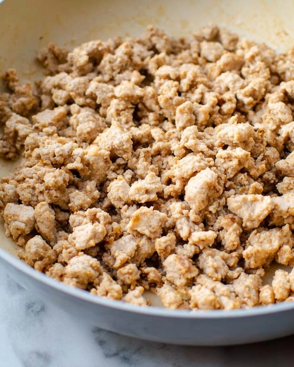 The image shows a close-up of many small, uneven pieces of cooked ground meat filling a white pan. The meat is light brown with some bits showing a slightly darker crisp texture, giving a crumbly look. The pan is sitting on a white marbled surface, and no other objects or colors are visible in the frame. The focus is on the texture and color of the cooked meat chunks. photo taken with an iphone --ar 4:5 --v 7