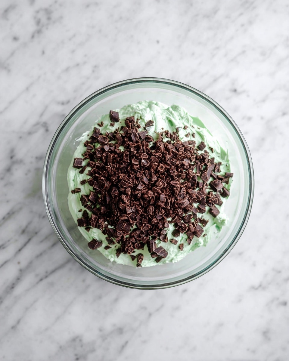 A clear glass bowl sits on a white marbled surface. Inside the bowl, there are two layers: the bottom layer is a soft, fluffy mint green cream, and the top layer is a generous pile of small dark brown chocolate pieces scattered unevenly over the green cream. The colors contrast sharply with each other, making both layers stand out clearly. photo taken with an iphone --ar 4:5 --v 7