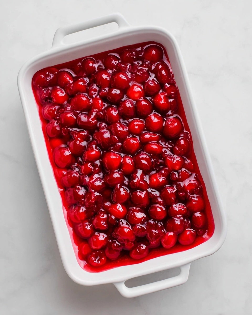 A white rectangular dish filled with one layer of bright red cherry topping that has a shiny, jelly-like texture and whole cherries visible throughout. The dish is set on a white marbled surface with the handles of the dish clearly visible on both sides. Photo taken with an iphone --ar 4:5 --v 7