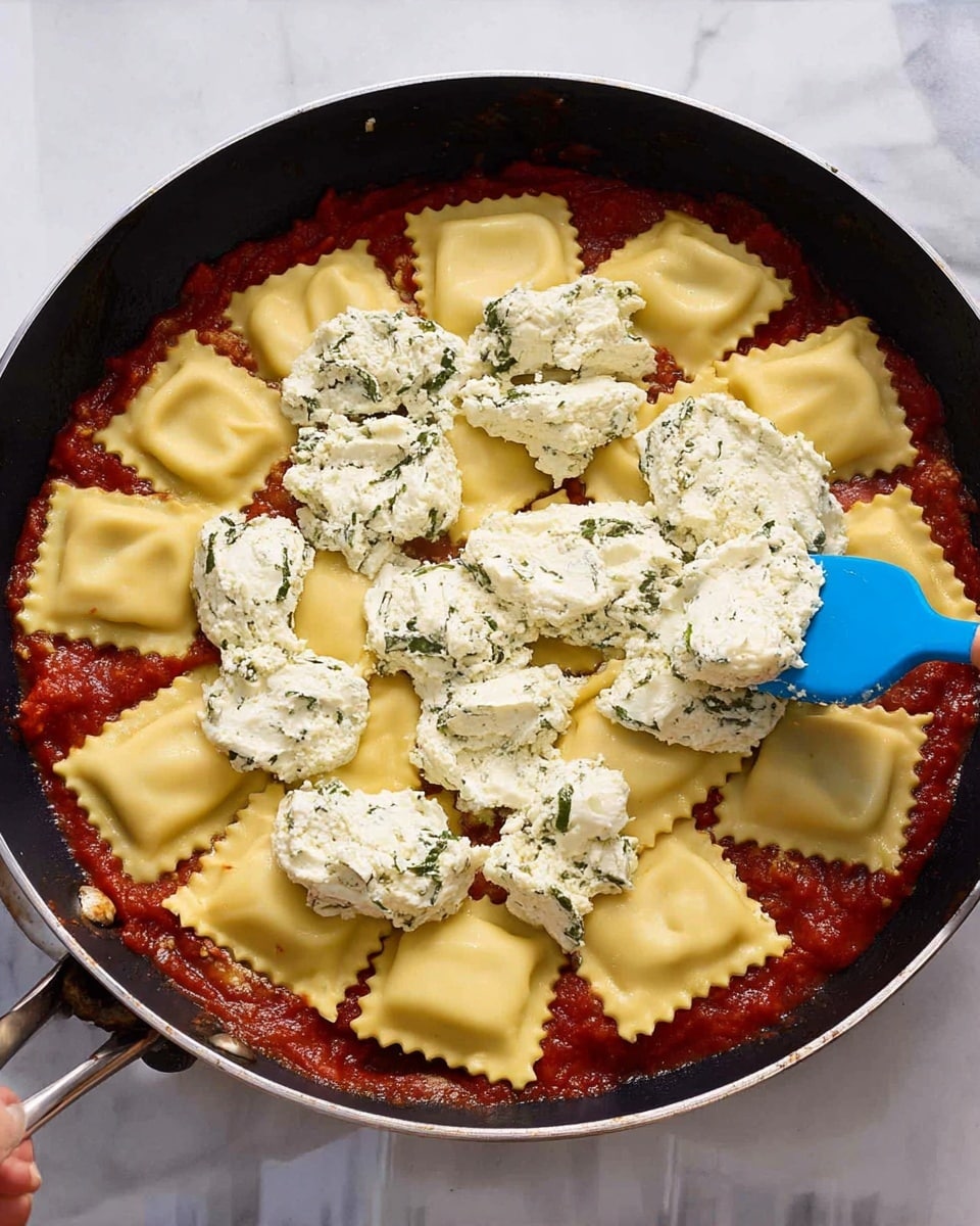 This image shows a black pan filled with three layers of food. The bottom layer is a red tomato sauce that spreads evenly across the pan, slightly thick and visible at the edges. The middle layer consists of large square ravioli pasta pieces placed in one even layer, showing a smooth, pale yellow surface and slightly puffed shape. On top, dollops of white ricotta cheese mixed with green herbs are spread unevenly over the ravioli. A woman's hand holding a blue spatula is placing some ricotta on the top right side of the pan. The pan rests on a white marbled surface. Photo taken with an iphone --ar 4:5 --v 7