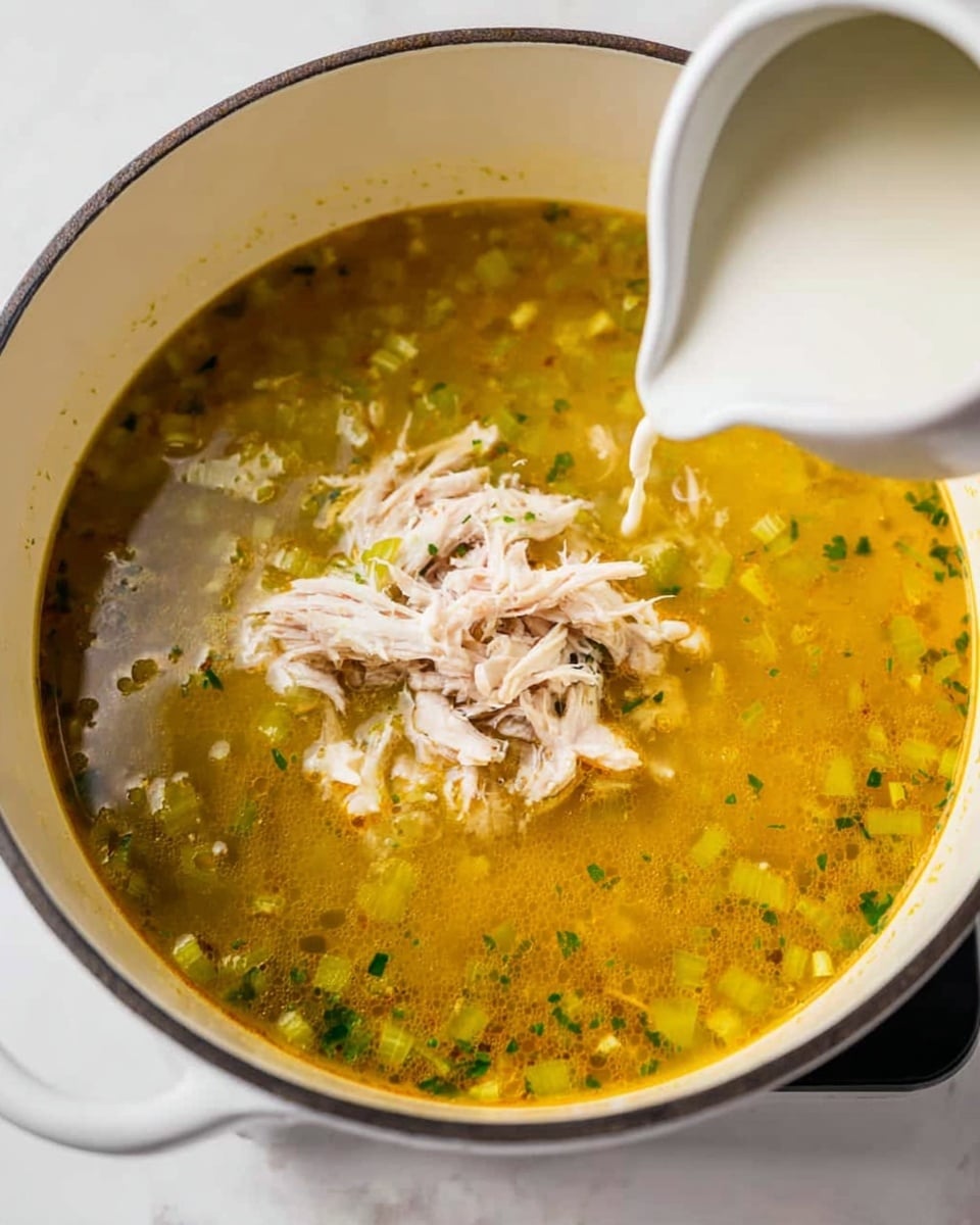 A pot of clear golden yellow soup filled with small pieces of green celery and herbs scattered throughout the broth. In the center, there is a small pile of shredded white chicken meat resting on top of the soup. A white cream is being poured into the pot from the right side by a woman's hand holding a small white pitcher. The pot sits on a white marbled surface, providing a clean and simple background. photo taken with an iphone --ar 4:5 --v 7