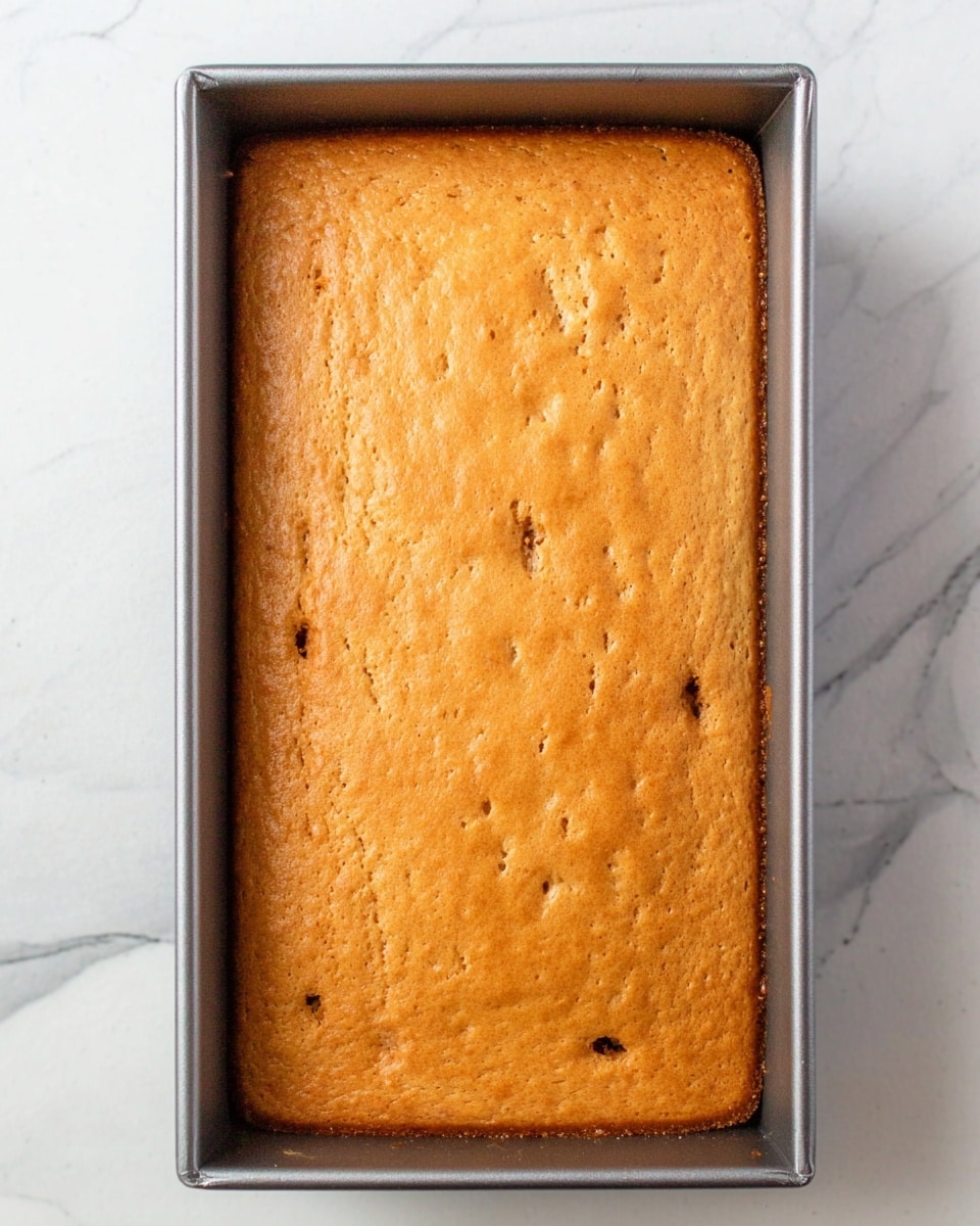 The image shows a full rectangular cake inside a metal baking tray placed on a white marbled surface. The cake has a smooth, golden brown top with small darker brown spots scattered across it, indicating texture and some crumbly parts. The edges of the cake are slightly darker than the center. The baking tray has a simple and clean look with straight edges, highlighting the soft, even surface of the baked cake. Photo taken with an iphone --ar 4:5 --v 7