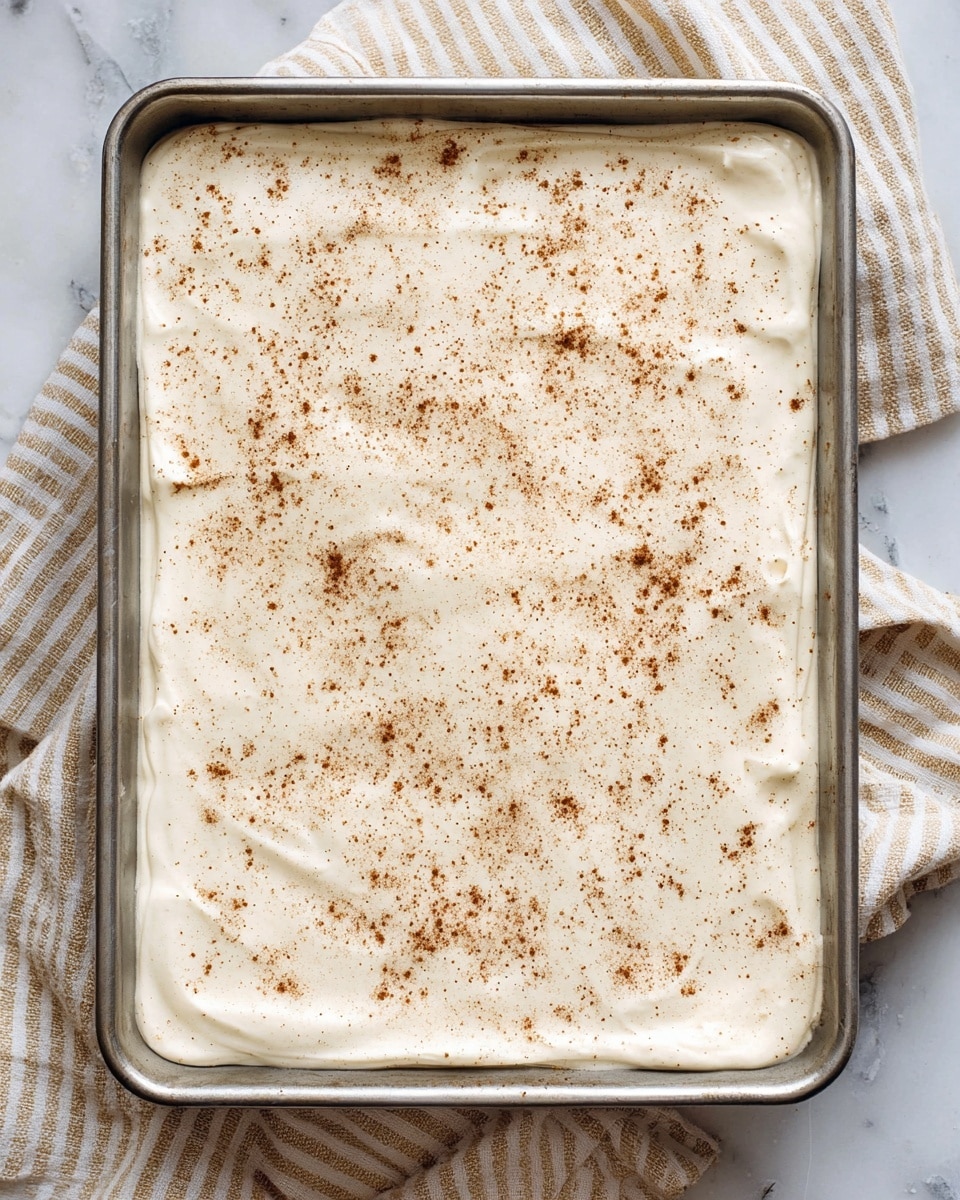 A rectangular metal pan filled with a smooth, thick white creamy layer evenly spread to the edges, topped with a light dusting of fine brown spice powder, likely cinnamon, scattered gently across the surface. The pan rests on a white marbled background with a beige and white striped cloth partially visible beneath it. The creamy layer looks soft and slightly textured, with small swirls and gentle peaks visible. photo taken with an iphone --ar 4:5 --v 7