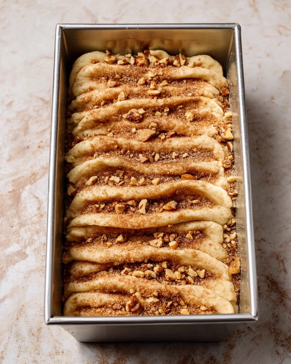 The image shows a rectangular metal baking pan filled with layers of dough and a cinnamon sugar mix sprinkled on top. The dough layers are placed close together, creating ridges that hold the light brown cinnamon sugar and small pieces of chopped nuts scattered evenly over the top. The baking pan is placed on a white marbled surface. Photo taken with an iphone --ar 4:5 --v 7
