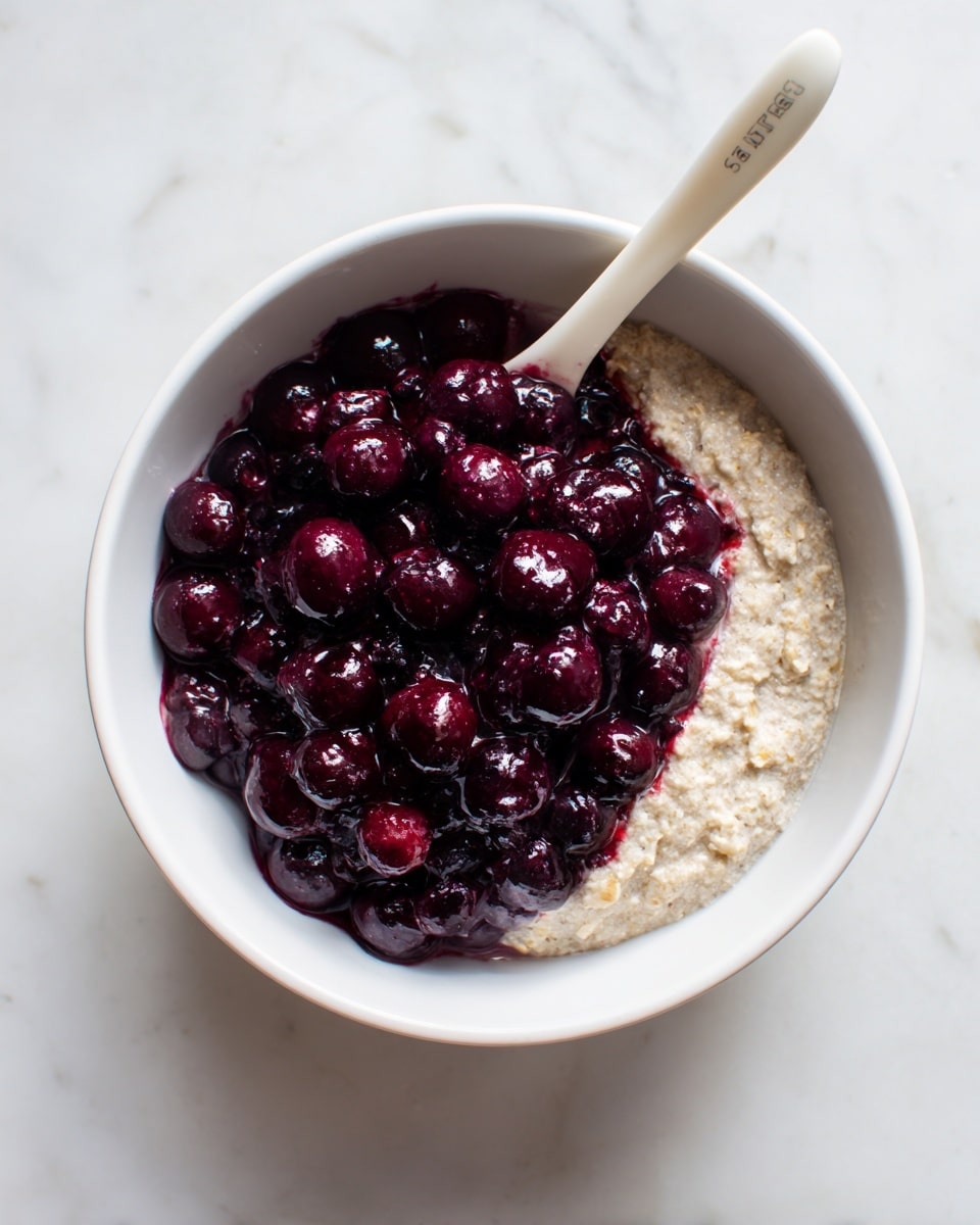 A white bowl contains two main layers: the bottom layer is a thick, light brown oat mixture with a slightly rough texture, filling about half the bowl, while the top layer is a generous pile of shiny, deep purple cooked berries, covering most of the oat layer but allowing a small part of it to show near the edge. A white plastic spoon with some text on the handle rests diagonally in the bowl, partially covered by the berries. The entire scene is set on a white marbled surface. Photo taken with an iphone --ar 4:5 --v 7