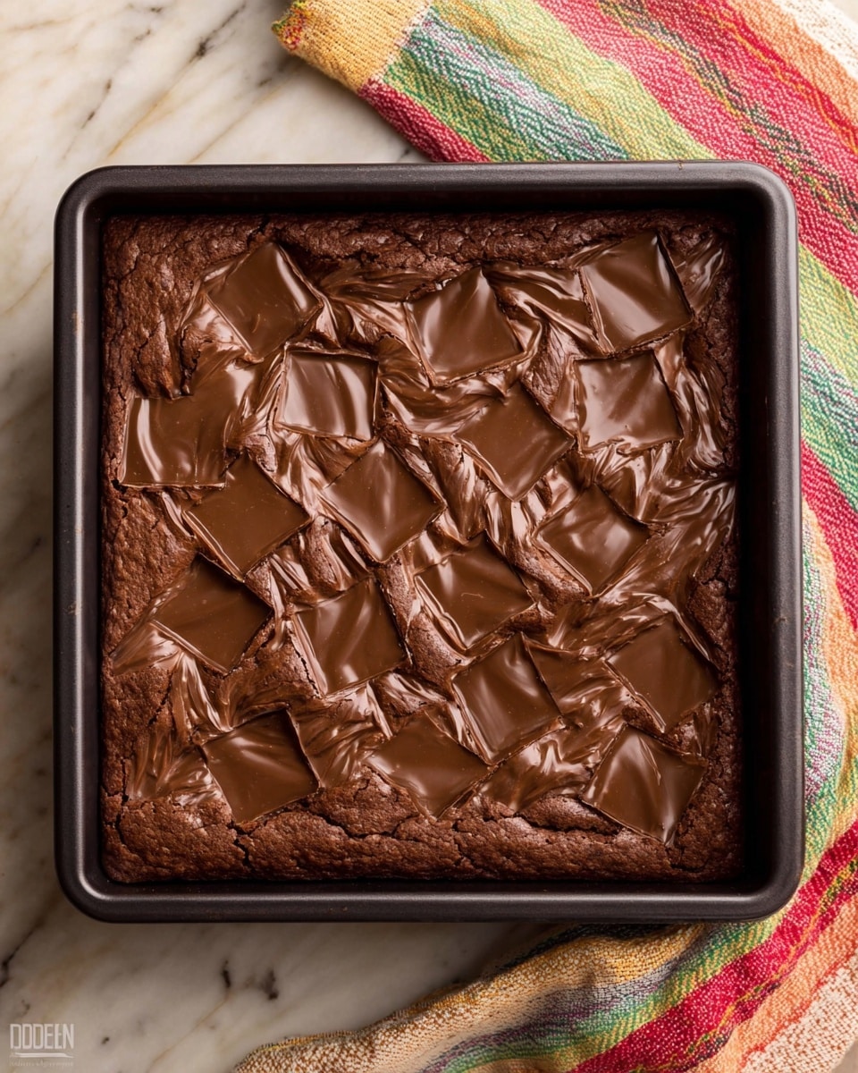A square brown baked chocolate dessert in a black baking pan, with many shiny square pieces of melted chocolate on top, evenly spread across the surface, showing a soft and slightly cracked texture. The background is a white marbled texture with a colorful striped cloth peeked in the bottom right corner. Photo taken with an iphone --ar 4:5 --v 7