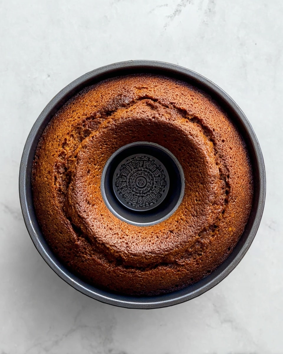 A single round bundt cake with a rich brown color sits in a dark metal bundt pan. The cake has a slightly rough texture on the top with uneven shades of darker and lighter brown, showing it is well baked. The cake's center hole reveals the patterned bottom of the pan. The pan is placed on a white marbled surface, giving a clean and bright background. photo taken with an iphone --ar 4:5 --v 7
