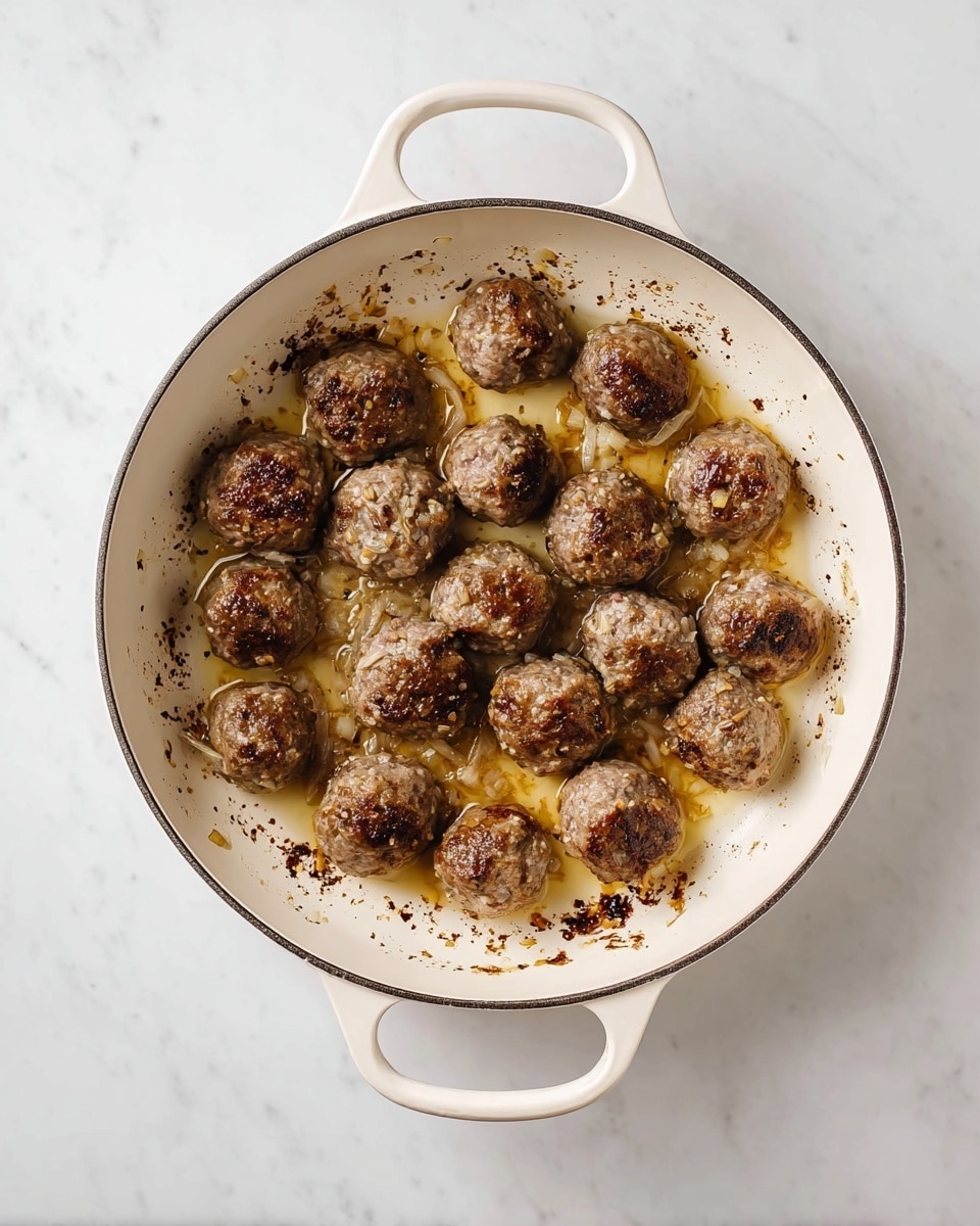 A white round pan with two handles contains thirteen small browned meatballs cooking in oil, scattered evenly. The meatballs have a rough texture with visible bits of onion. The pan shows brown spots of cooked onion and oil glistening around the meatballs. The pan is set on a white marbled surface. Photo taken with an iphone --ar 4:5 --v 7