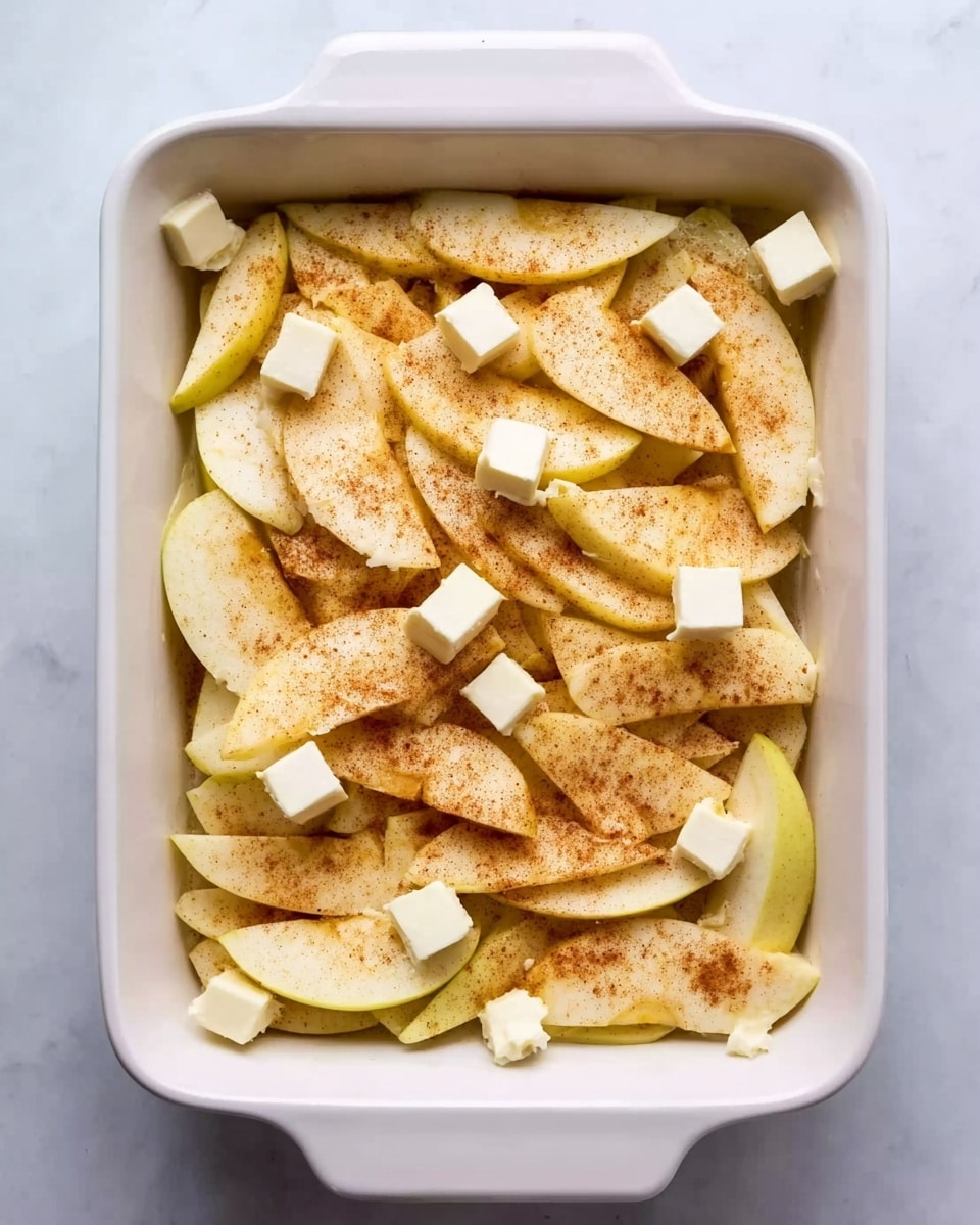 The image shows a white rectangular baking dish filled with thin apple slices spread evenly in one layer. The apple slices are pale yellow with a light dusting of cinnamon or brownish spice. Small cubes of butter are scattered on top of the apple slices, adding a creamy white contrast. The dish sits on a white marbled surface, giving a clean and bright background. Photo taken with an iphone --ar 4:5 --v 7