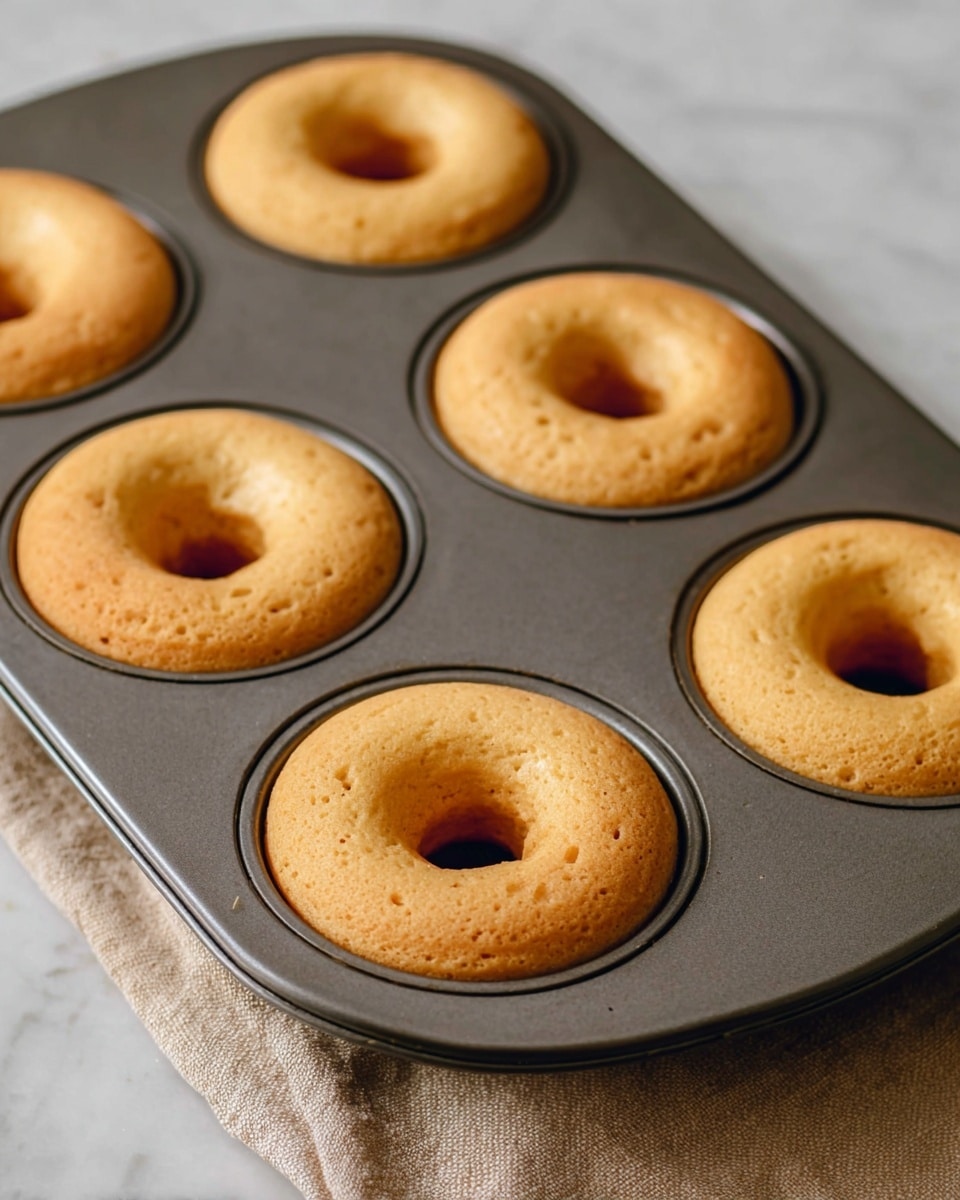 The image shows six freshly baked plain donuts in a dark gray metal donut baking tray. Each donut has one light golden brown layer with a smooth and slightly raised top texture, showing small bubbles on the surface. The donuts fit neatly inside the round molds of the tray, and the view is close-up from the side angle. The tray sits on a light brown cloth, and both are placed on a white marbled surface. Photo taken with an iphone --ar 4:5 --v 7