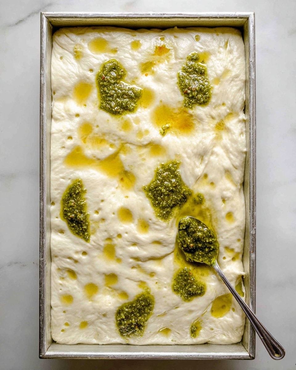 A thick, fluffy white dough fills a silver rectangular baking pan, showing small air bubbles and dimples on its surface. There are several uneven dollops of bright green pesto sauce scattered on top, with a spoon near the bottom right corner spreading more pesto over the dough. The dough has some shiny golden spots where the oil from the pesto has soaked in. The setting is on a white marbled surface. photo taken with an iphone --ar 4:5 --v 7