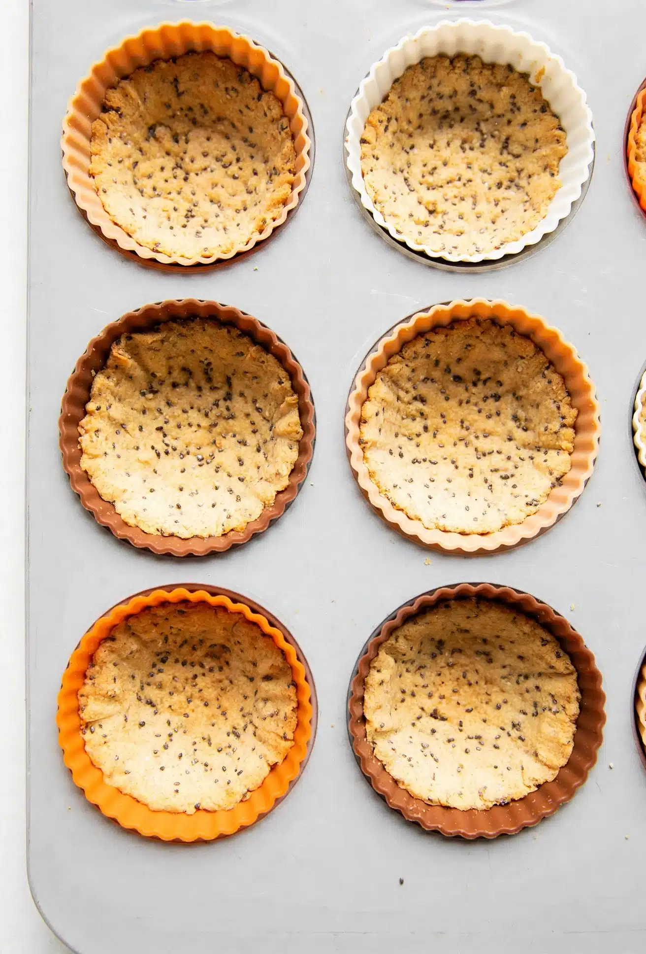 American Flag Fruit Tarts Recipe 5 The image shows eight tart pans arranged in two vertical columns and four horizontal rows on a white marbled surface. Each round tart pan is filled with dough containing visible small black seeds mixed evenly throughout. The pans come in different colors: orange, white, and brown, with the dough pressed inside each to form a uniform base layer about two to three centimeters thick. The dough texture looks smooth with some light indentations. The pans’ ridged edges are clearly visible, creating a scalloped pattern around each tart shell. The overall scene is well-lit, giving a clean and fresh impression. Photo taken with an iphone --ar 4:5 --v 7
