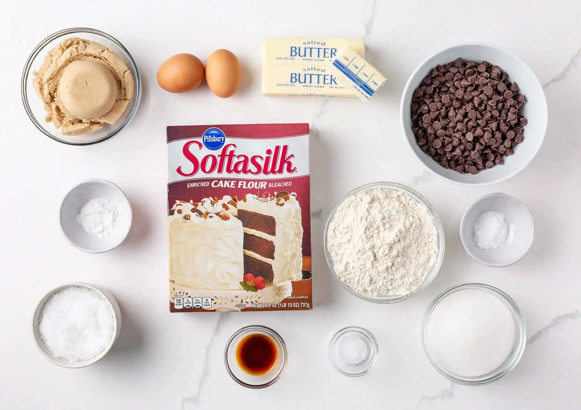 Jacques Torres Chocolate Chip Cookies Recipe 4 The image shows various baking ingredients arranged neatly on a white marbled surface. In the center, there is a box of Pillsbury Softasilk cake flour with a picture of a layered white cake with chocolate layers and cream frosting on the box. Surrounding the box are glass bowls and white bowls with different ingredients: light brown sugar in a glass bowl, two brown eggs, two sticks of salted butter, a white bowl filled with chocolate chips, a bowl with white flour, a bowl with white granulated sugar, a small white bowl with baking powder, a tiny clear bowl with salt, a small bowl with white powdered sugar, and a tiny glass bowl holding vanilla extract. The ingredients are spaced evenly and organized simply. Photo taken with an iphone --ar 4:5 --v 7
