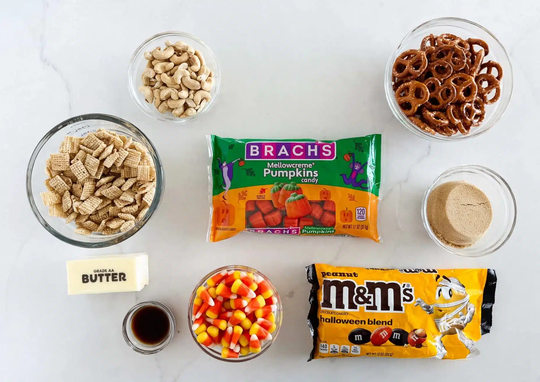 Fall Chex Mix Recipe 4 The image shows a flat lay of nine ingredients arranged neatly on a white marbled surface. There is a clear glass bowl with light beige square cereal pieces in the bottom left, next to a stick of pale yellow butter labeled