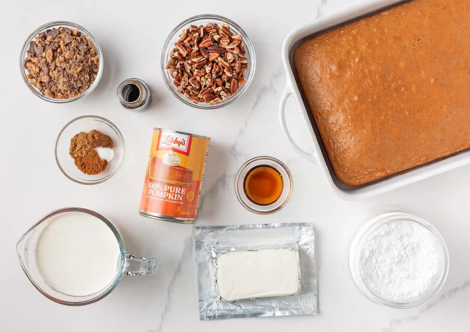 Pumpkin Trifle with Spice Cake, Cream Cheese, and Toffee Recipe 4 A white baking dish with a smooth, golden brown pumpkin cake filling most of the frame's right side shows a flat baked layer with a slightly textured top. On the left side, arranged on a white marbled surface, are several clear glass bowls with ingredients: one bowl has small brown chunks, another bowl with whole pecan halves, a small round bowl with light brown, dark brown, and white powders, and a larger measuring pitcher filled with white cream. Near the center are a small glass bowl with amber liquid, an open tin of pure pumpkin puree, a block of cream cheese on foil, and a white jar filled with powdered sugar. The scene is bright and clean, photo taken with an iphone --ar 4:5 --v 7