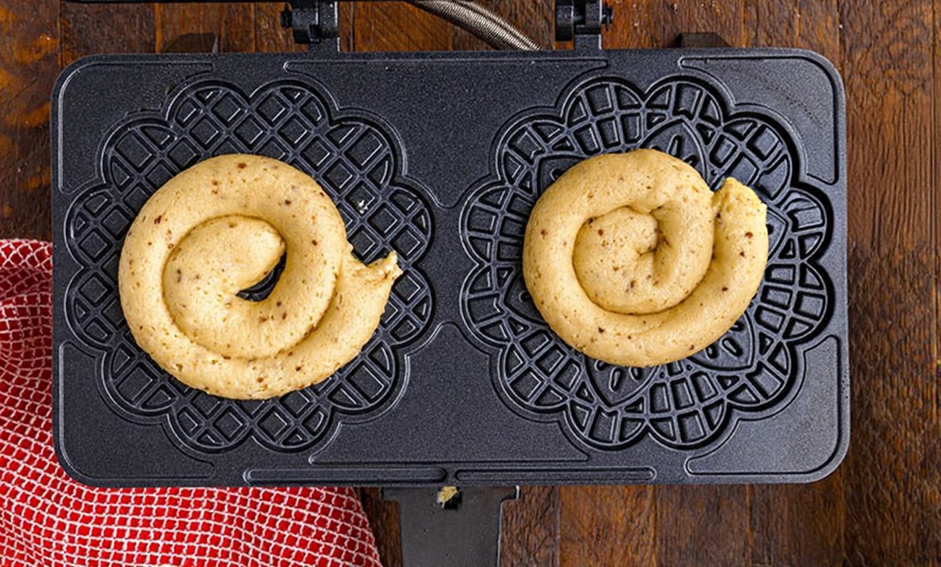 The image shows a black waffle iron with two heart-shaped molds filled with light beige batter that has small darker brown specks. The batter is swirled in a spiral shape in each mold, with the one on the left being slightly larger than the one on the right. The waffle iron rests on a wooden surface, and a red and white textured cloth is placed to the left side. The surface is changed to a white marbled texture behind the setup. photo taken with an iphone --ar 4:5 --v 7