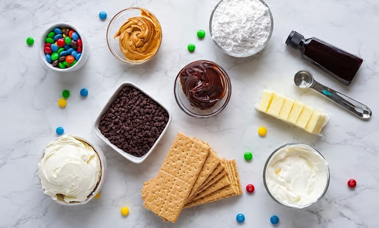 The image shows a flat top-down view of various ingredients arranged on a white marbled surface. There is a bowl of colorful candy-coated chocolates in green, blue, red, yellow, and brown, a bowl of smooth brown peanut butter, and a glass bowl filled with powdered sugar. Next to them are a small glass bowl of dark chocolate spread and a silver measuring spoon near a bottle of dark liquid, possibly vanilla extract. Several sticks of butter are neatly sliced and placed in a row, with a small bowl of creamy white frosting nearby. A square white dish holds mini chocolate chips, and several rectangular graham crackers are stacked in the center. Candy-coated chocolates are scattered around the surface, adding bright pops of color. photo taken with an iphone --ar 4:5 --v 7