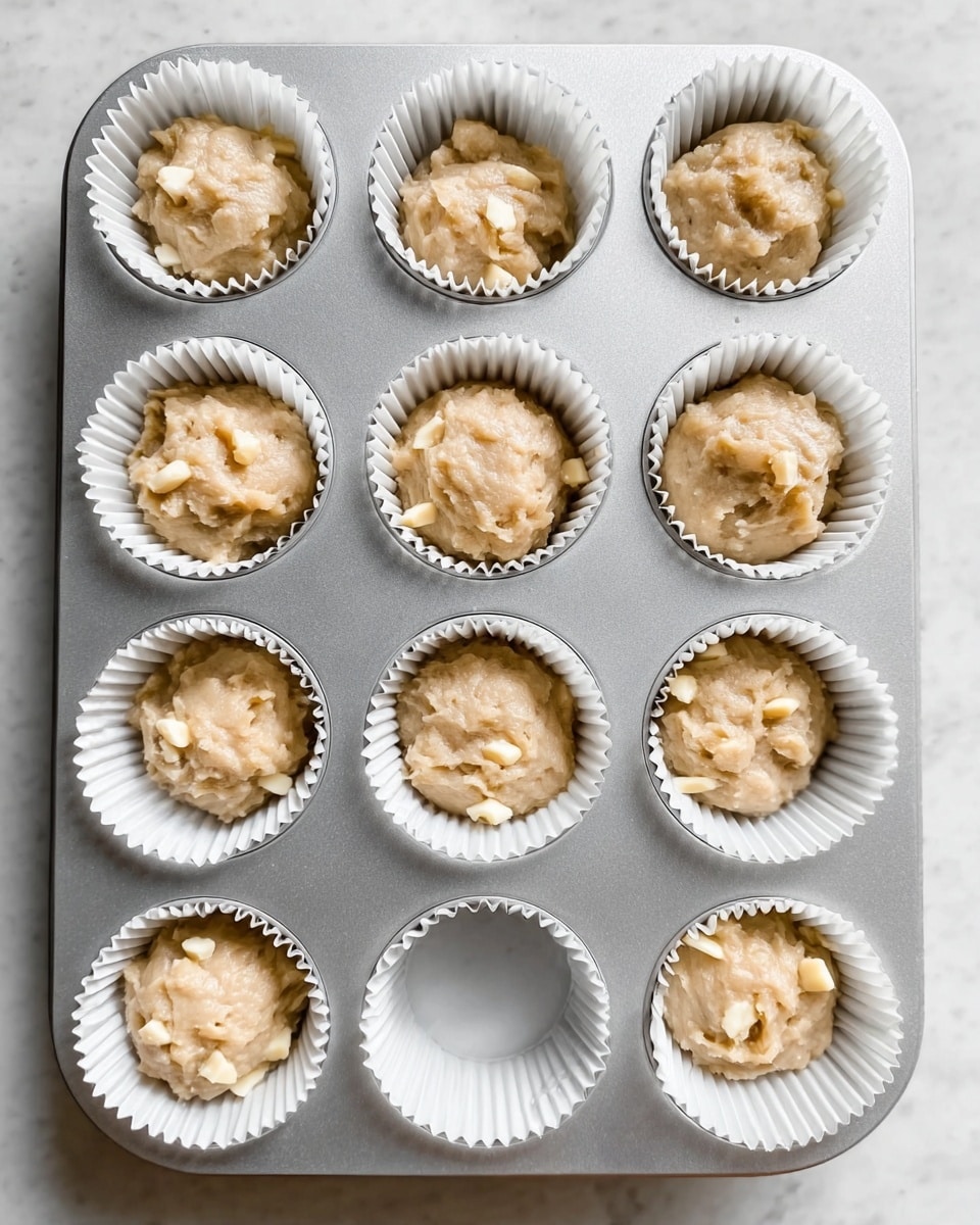 Nine white paper-lined muffin slots in a silver metal tray each hold a scoop of light beige muffin batter with visible small pale yellow chunks mixed inside. The batter has a soft, slightly rough texture, looking dense but moist. Three empty white paper liners are at the bottom of the tray. The background is a white marbled surface, and the photo is taken from above, showing a clean and bright kitchen setting. Photo taken with an iphone --ar 4:5 --v 7