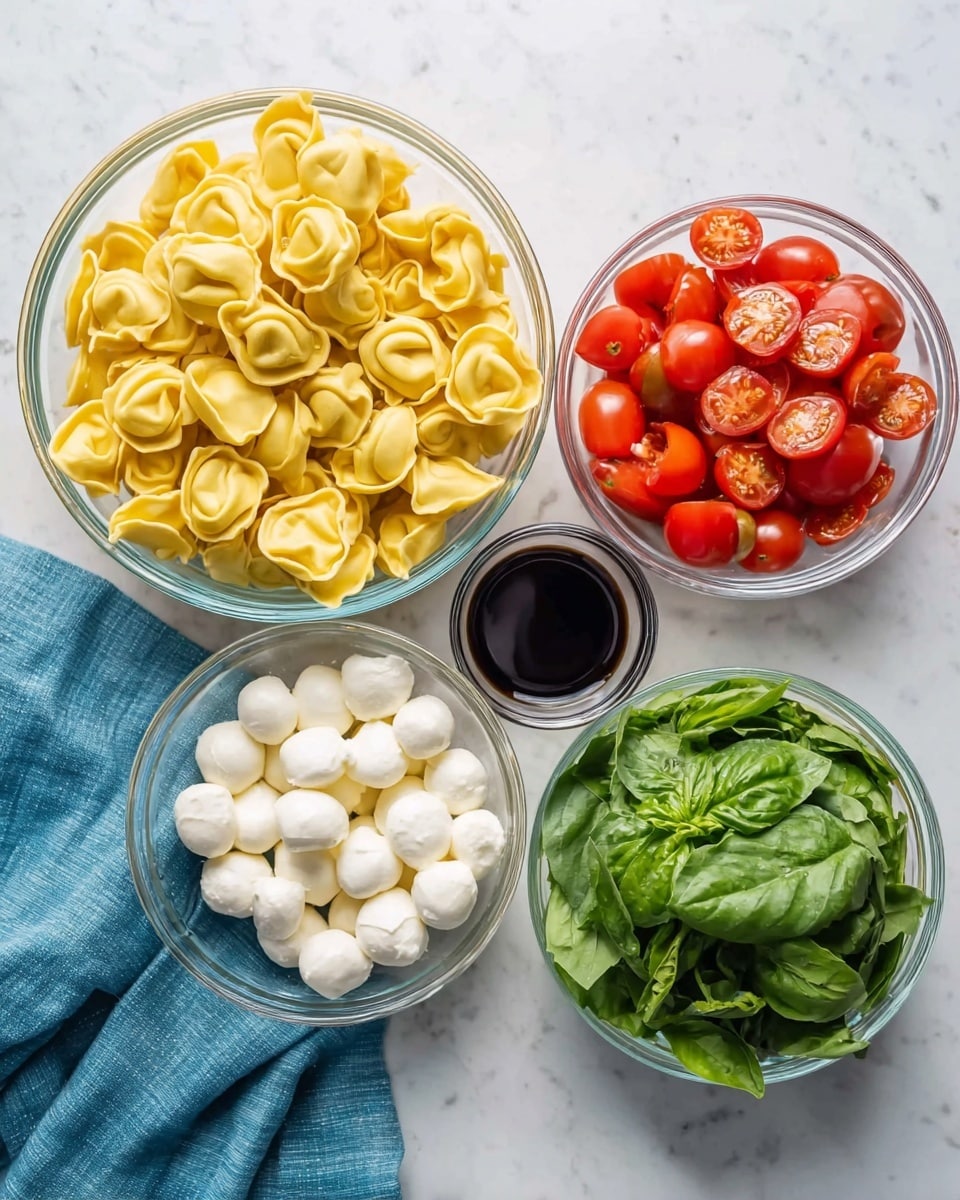 The image shows five glass bowls arranged on a white marbled surface with a blue cloth under the left bowl. The largest bowl on the left is filled with uncooked yellow tortellini pasta, showing their folded and stuffed texture. The bowl on the top right contains a mix of bright red, halved grape tomatoes on one side and fresh green spinach leaves on the other. Below that is a smaller bowl filled with small white mozzarella balls. To the left of the mozzarella is a small bowl with dark balsamic vinegar or soy sauce, smooth and shiny. The smallest bowl at the bottom holds fresh green basil leaves with a soft texture. Photo taken with an iphone --ar 4:5 --v 7