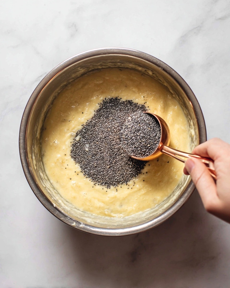 Inside a metal mixing bowl sits a thick, pale yellow batter with a soft and slightly lumpy texture. A woman's hand is holding a small copper measuring cup, pouring a heap of small, round, dark grey poppy seeds onto the center of the batter. The bowl is placed on a white marbled surface which serves as the background. The batter fills about half of the bowl, and the poppy seeds create a rough, grainy layer on top, centered and contrasting with the smoothness of the batter below. photo taken with an iphone --ar 4:5 --v 7