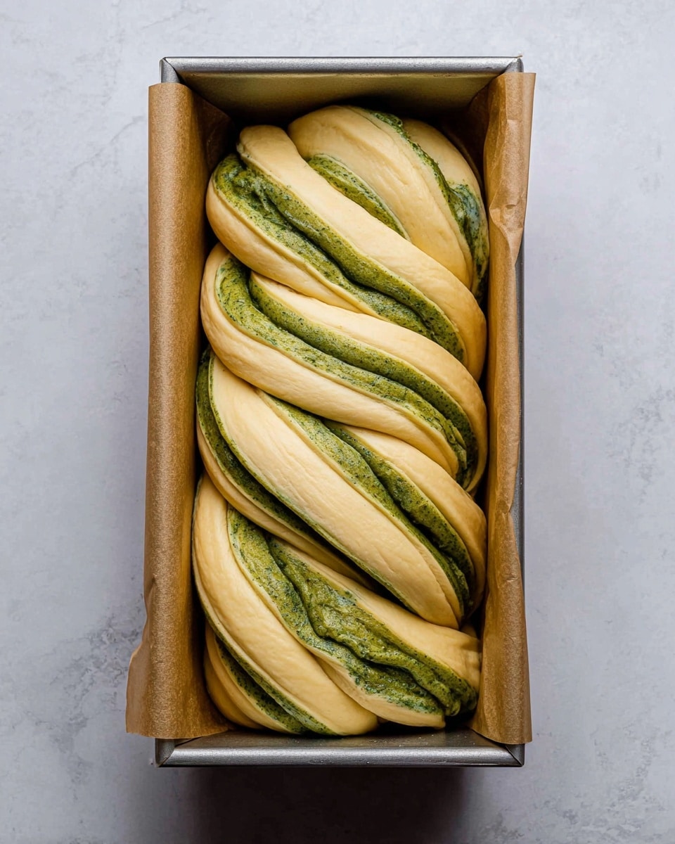 The image shows a twisted dough loaf inside a rectangular metal baking pan lined with brown parchment paper. The dough has visible layers, alternating between a light beige dough and a green filling that runs through each layer, creating a swirling pattern. The twists are thick, with the green filling forming wavy streaks within the dough, giving it a textured look. The pan rests on a white marbled surface. photo taken with an iphone --ar 4:5 --v 7
